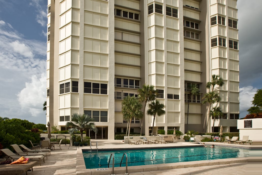 Condominium tower behind a swimming pool with palm trees and lounge chairs.