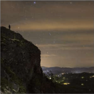 Rocky mountain cliff at night overlooking distant hills dotted with town lights.