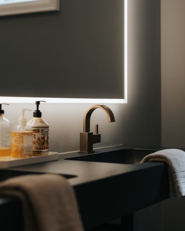 Bathroom vanity with an arched brass faucet, backlit mirror, soap bottles, and towels.