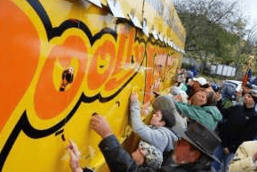 Crowd reaching toward a brightly painted yellow surface at an outdoor event.