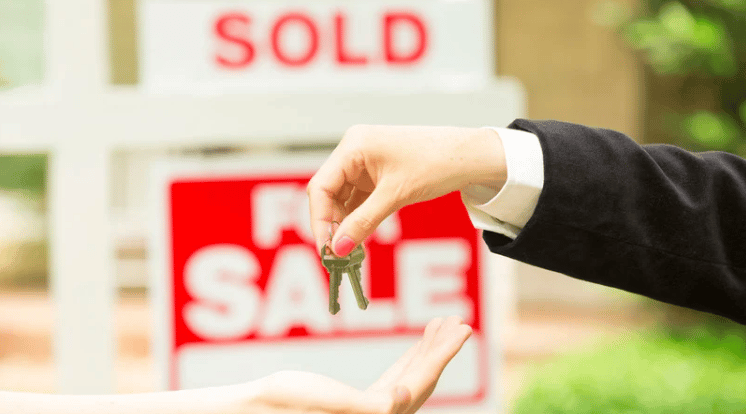 House keys change hands in front of a For Sale sign and a blurred Sold sign.