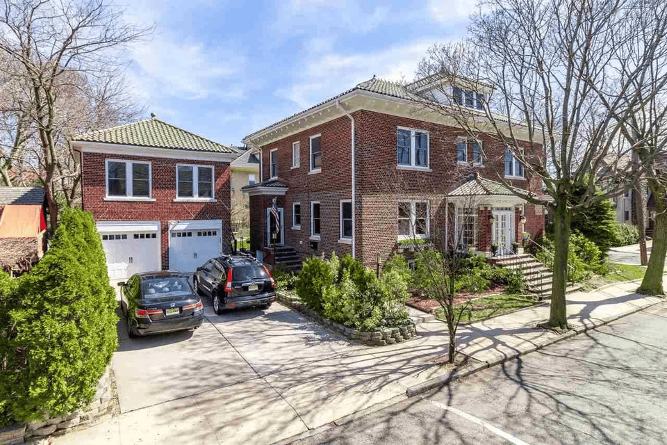 Two-story brick residence with adjoining garage wing, driveway, parked cars, and landscaped shrubs.