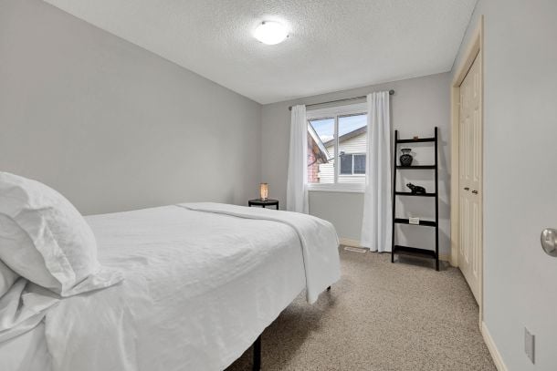 Bedroom with crisp white bedding, beige carpet, and a window with white curtains.