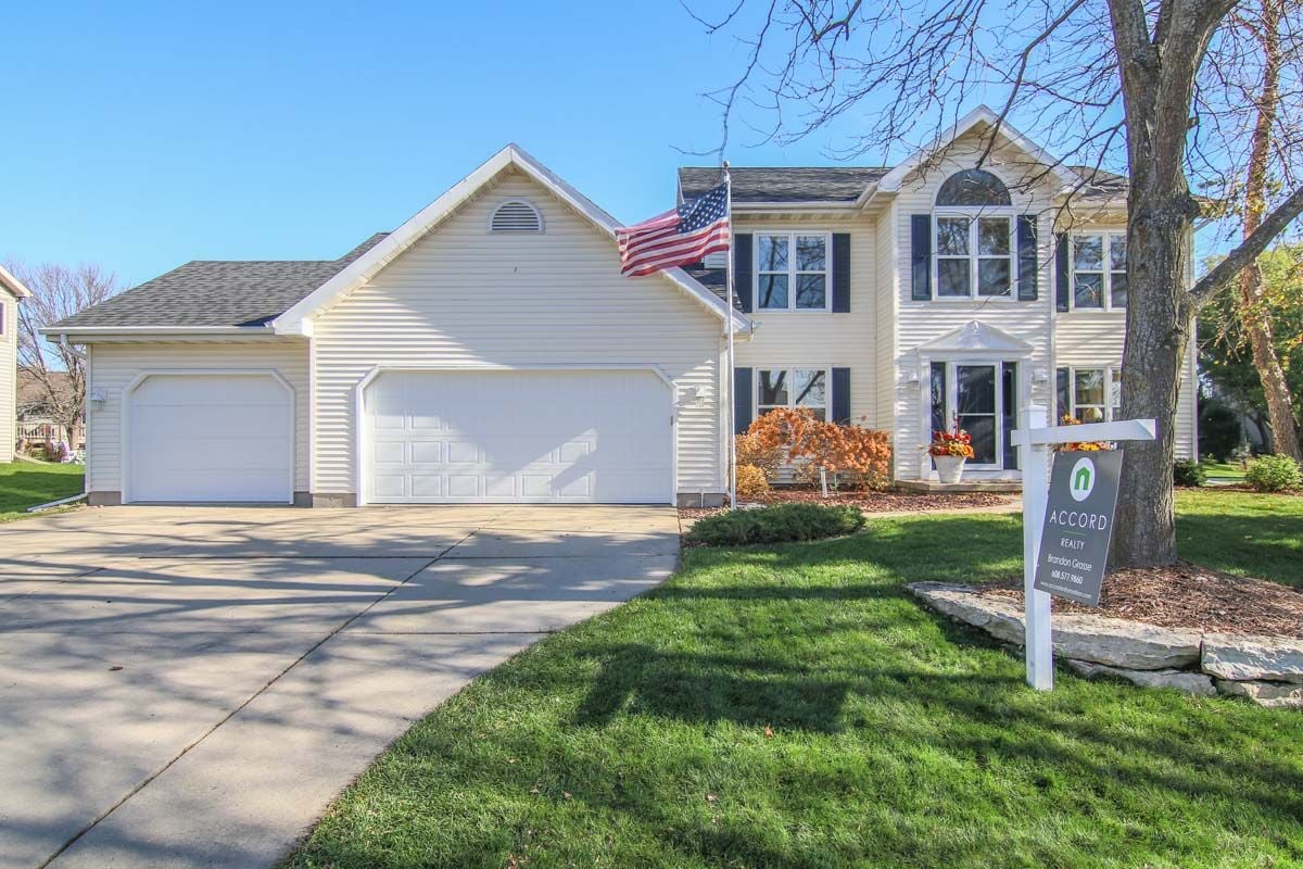White colonial home with attached garages, an American flag, and a realty sign by the lawn.