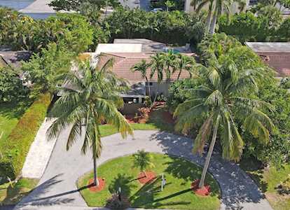 Aerial view of a home with a curved driveway, palms, and landscaped lawn.