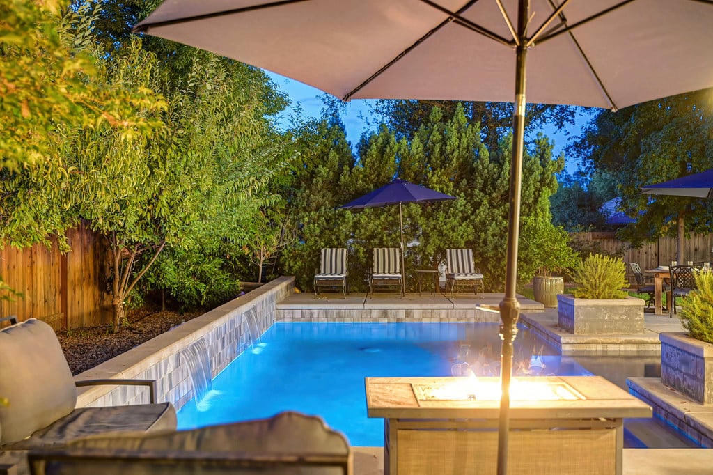 Backyard swimming pool at dusk with stone coping, patio seating, and a glowing fire table.