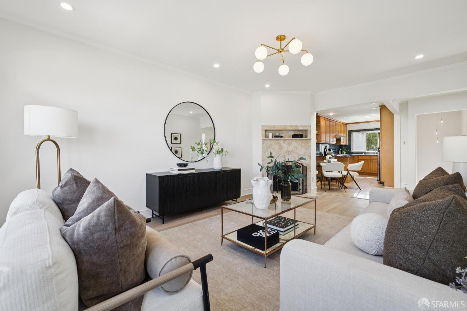 Staged living room with a stone fireplace, round mirror, and open view into the wood-cabinet kitchen.
