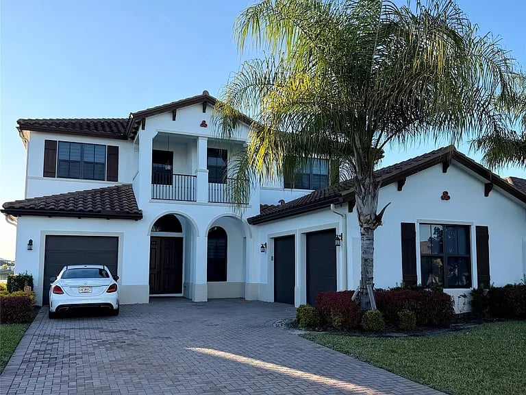 Two-story white stucco home with arched entryways, dark tiled roof, and a palm tree by the driveway.