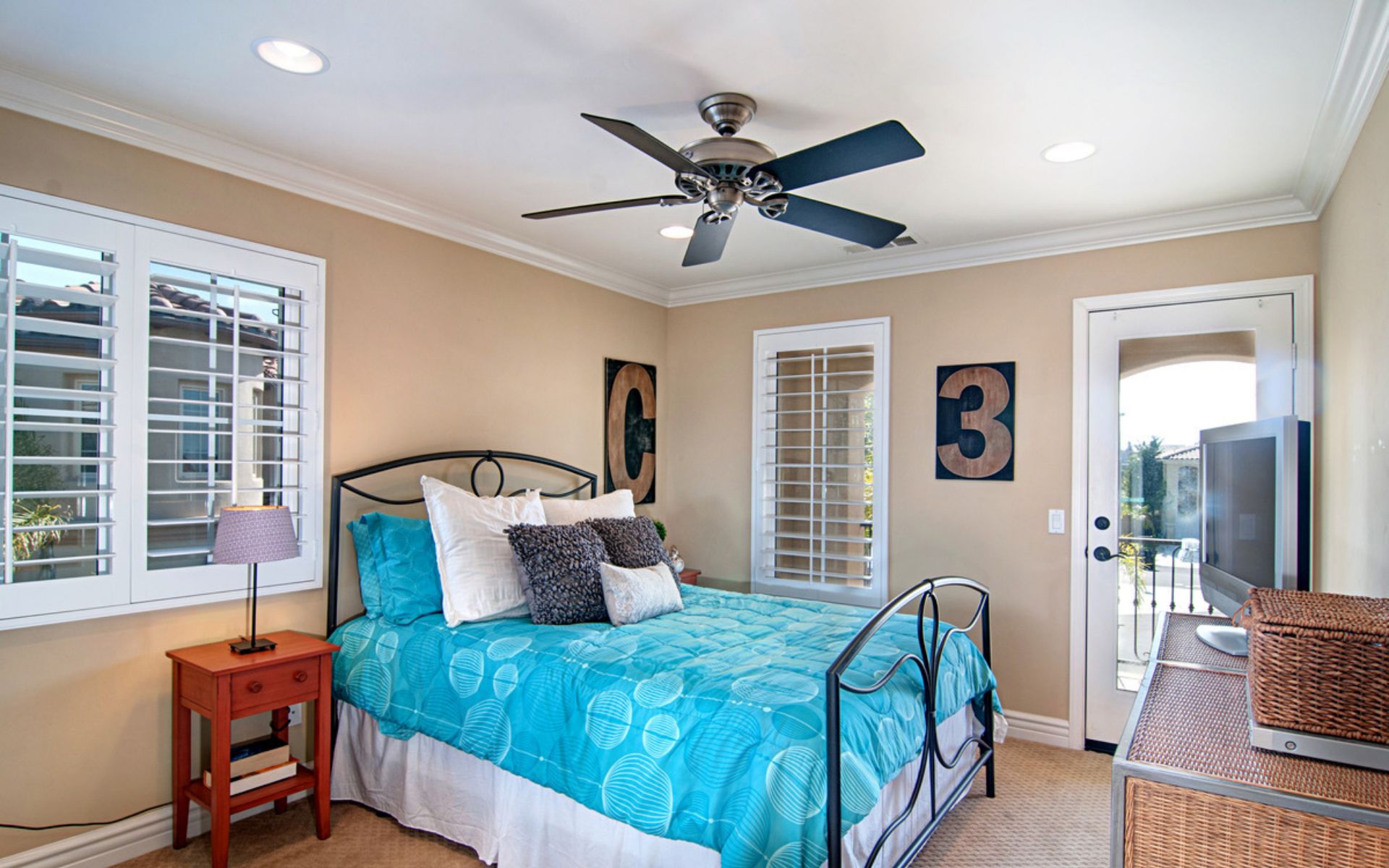 Bedroom with a turquoise patterned bedspread, black metal bed frame, plantation shutters, and ceiling fan.
