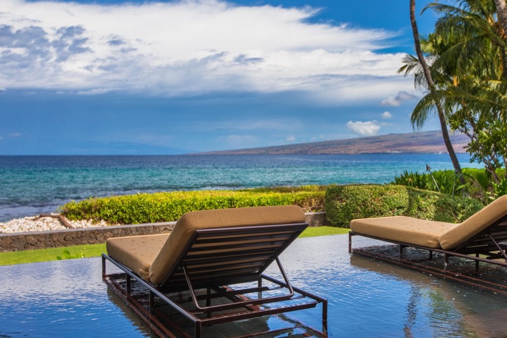 Poolside loungers overlook turquoise ocean water, a distant coastline, and palm trees.