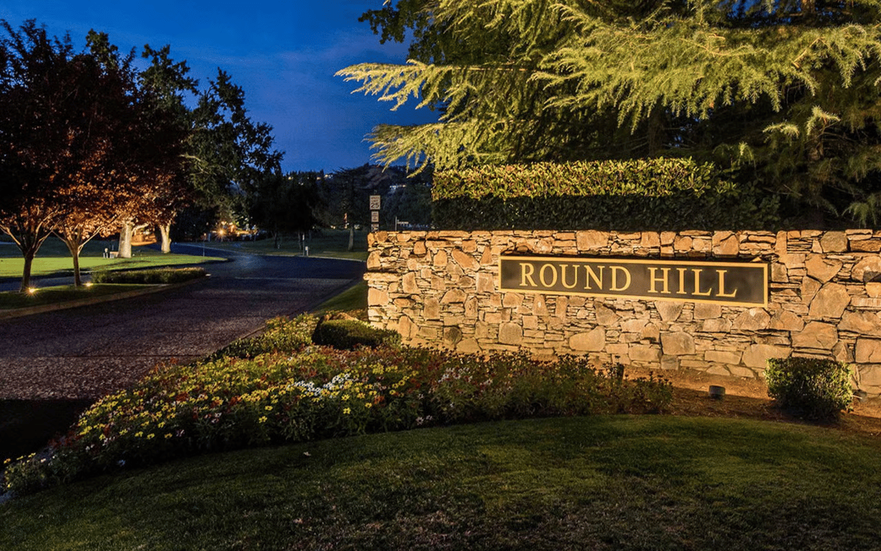 Round Hill entrance sign on a stone wall, illuminated beside a landscaped road at night.