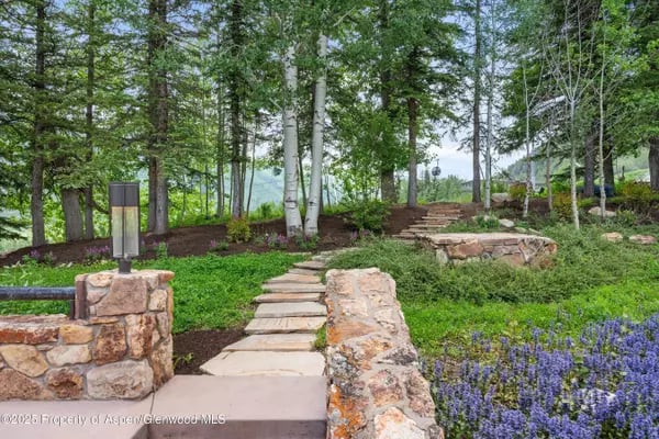 Stone walkway and steps bordered by low stone walls, mature trees, and purple flowers.