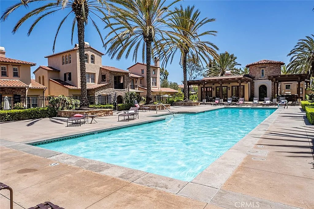 Outdoor swimming pool with palm trees, lounge chairs, and Mediterranean-style buildings.