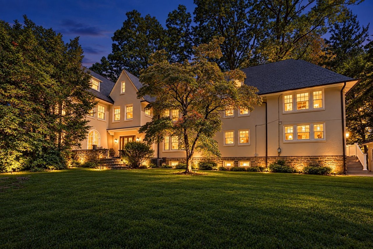 Two-story home with warmly lit windows, stone foundation, and mature trees at twilight.