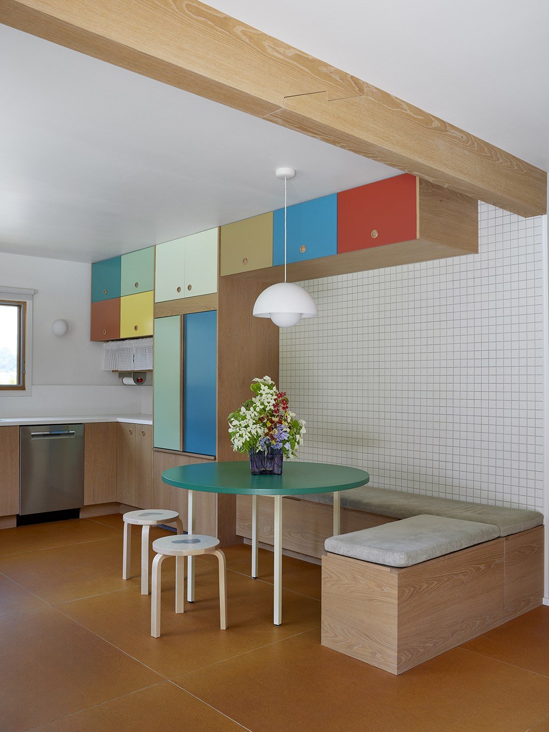 A kitchen with color-blocked upper cabinets in pale green, blue, yellow and red, under an exposed wood beam, with a round green dining table.