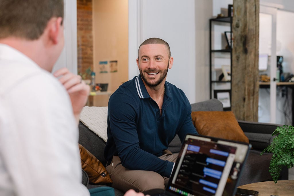 A smiling man discusses home buying with another person beside an open laptop.