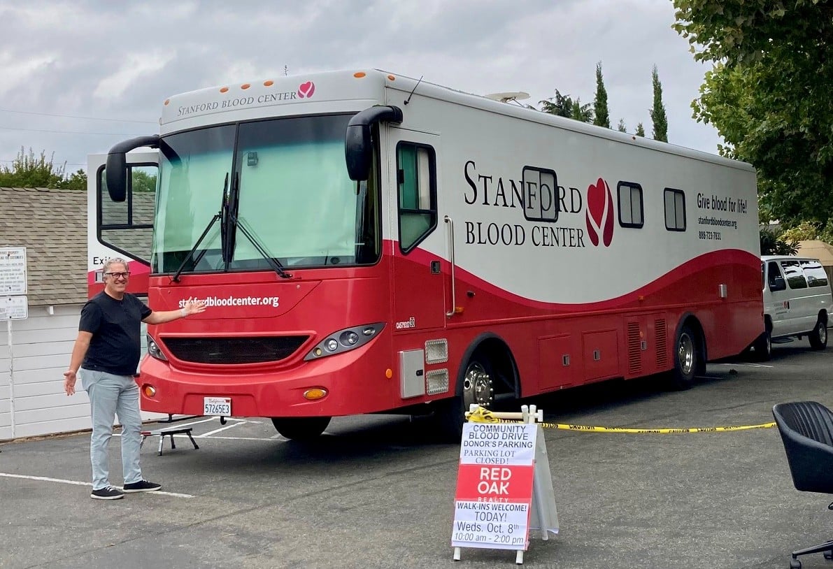 A man welcomes visitors beside a Stanford Blood Center blood-drive bus and community parking sign.