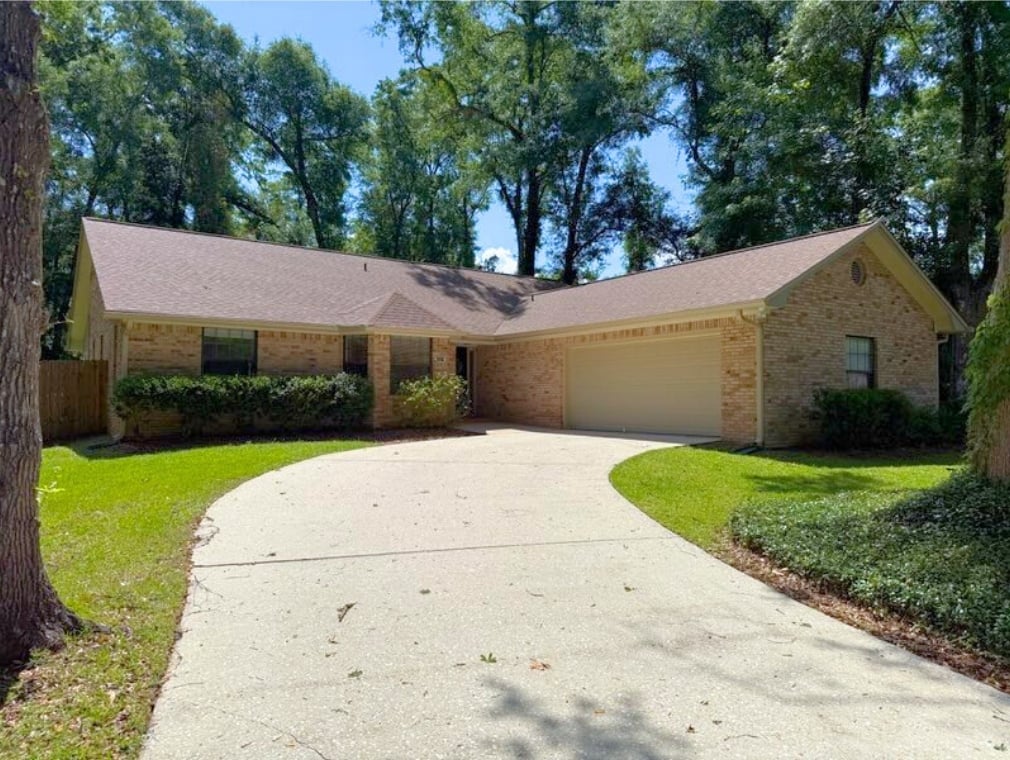 A one-story brick house with a brown roof sits in a wooded area. A curved driveway leads to the garage, surrounded by neatly trimmed bushes and green grass.
