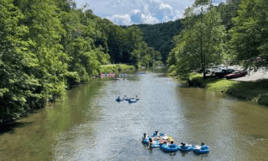 People tubing on a tree-lined river, with wooded hills and boats along the bank.