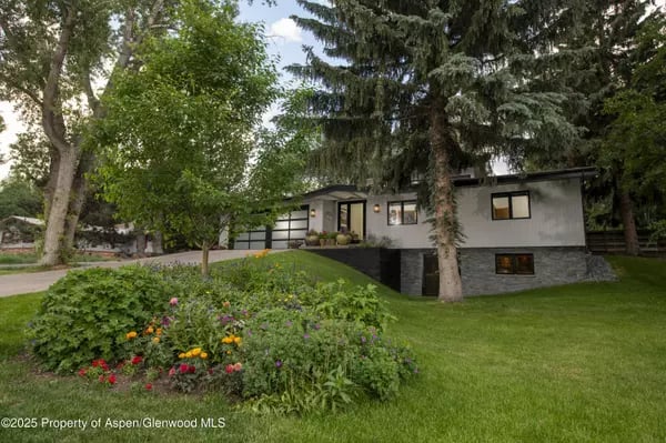 Contemporary home with a dark stone facade, mature trees, and a colorful flower garden.