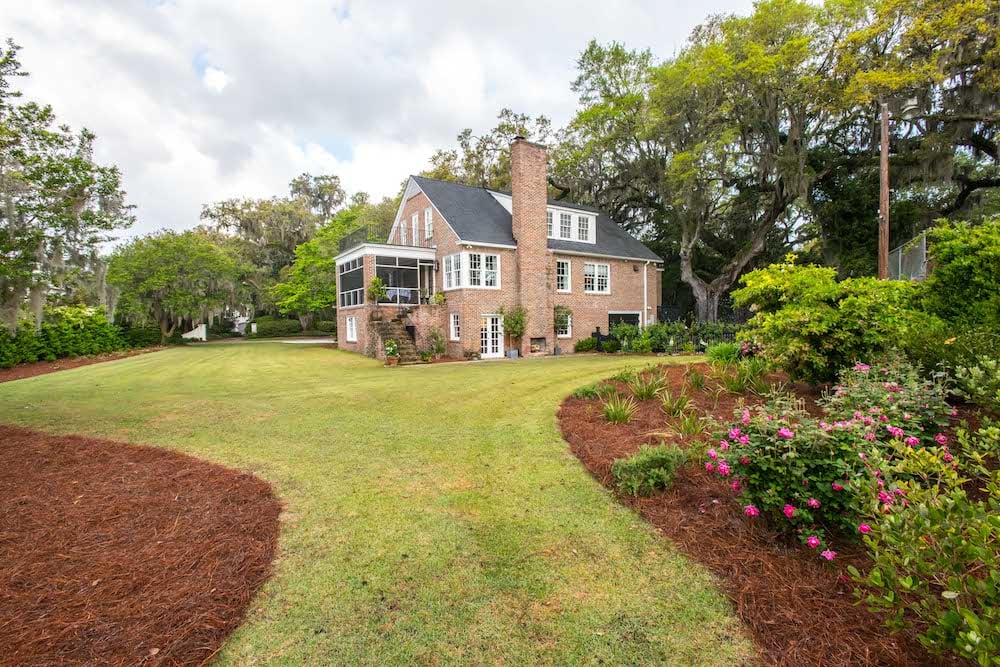 Brick house beyond a broad lawn, with curved garden beds and flowering shrubs.