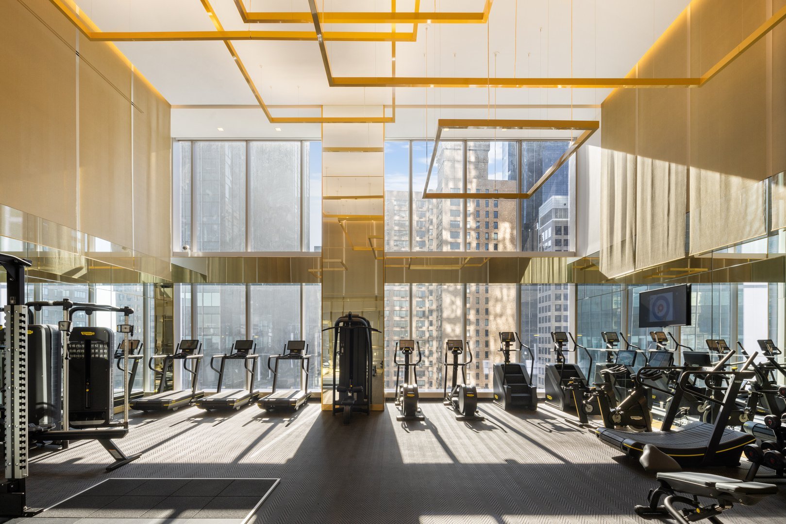 High-rise fitness center with rows of exercise machines, floor-to-ceiling windows, and sunlight.