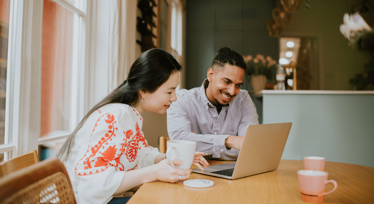A couple smiles while looking at a laptop together at a wooden table.