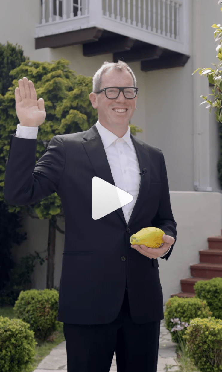 A suited man waves while holding a yellow fruit in front of a home.