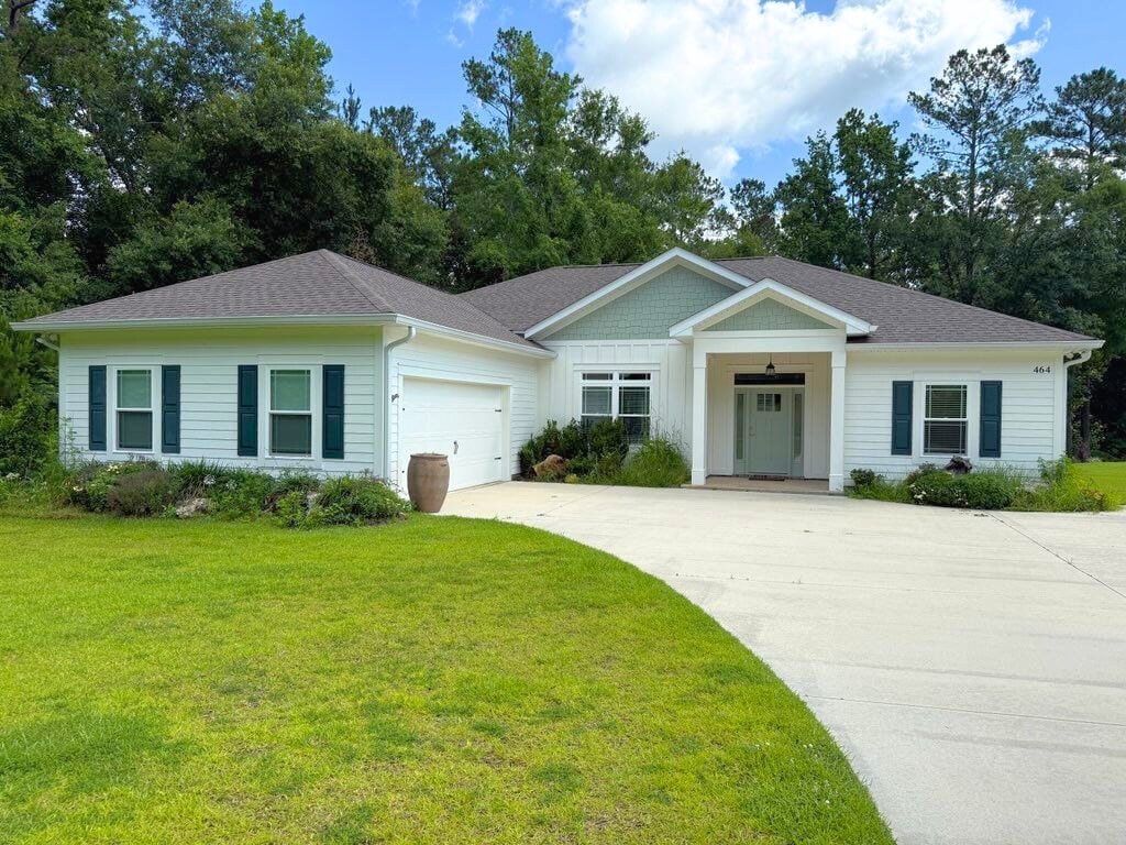 Single-story house with light blue siding and a dark roof. It has a large driveway, green lawn, and a wooded background, conveying a serene suburban feel.