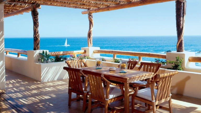 Oceanfront terrace with a wooden dining set beneath a timber pergola and a sailboat offshore.
