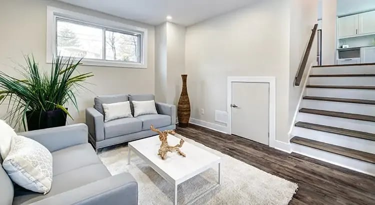 A spacious, modern living room with light-colored furniture, a large rug, and a staircase leading to another level. The room is well-lit with natural light.