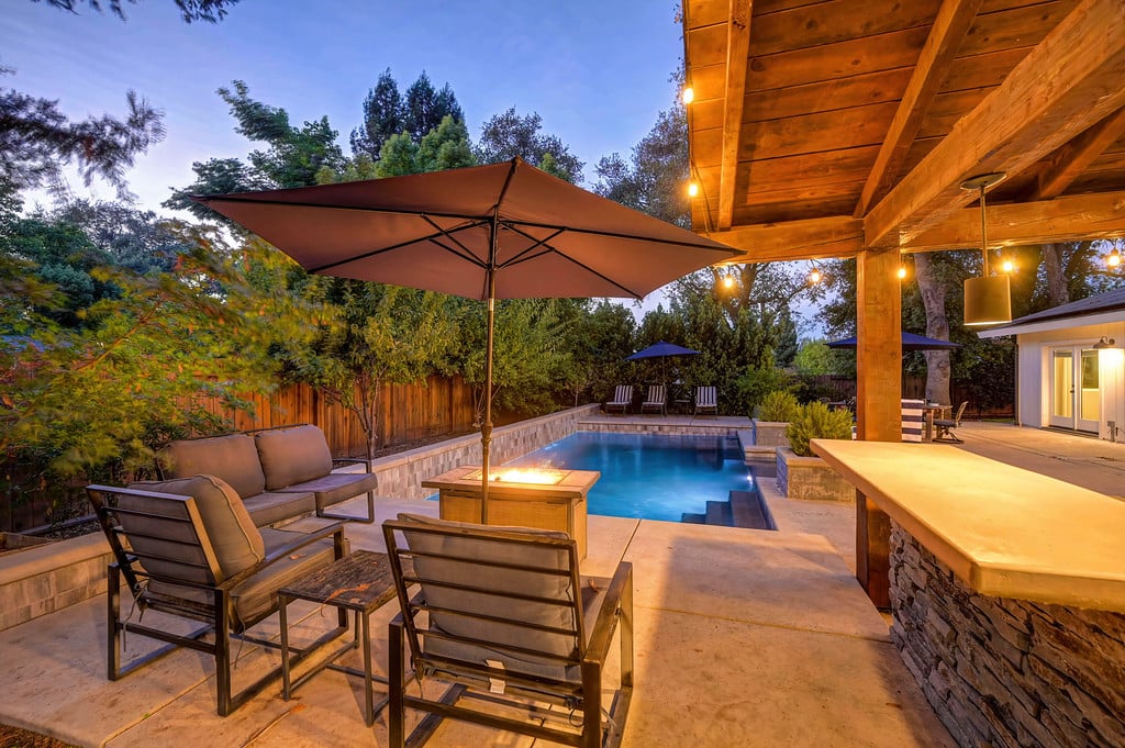 Backyard pool and covered patio with lounge chairs, an umbrella, and a lit outdoor bar at dusk.