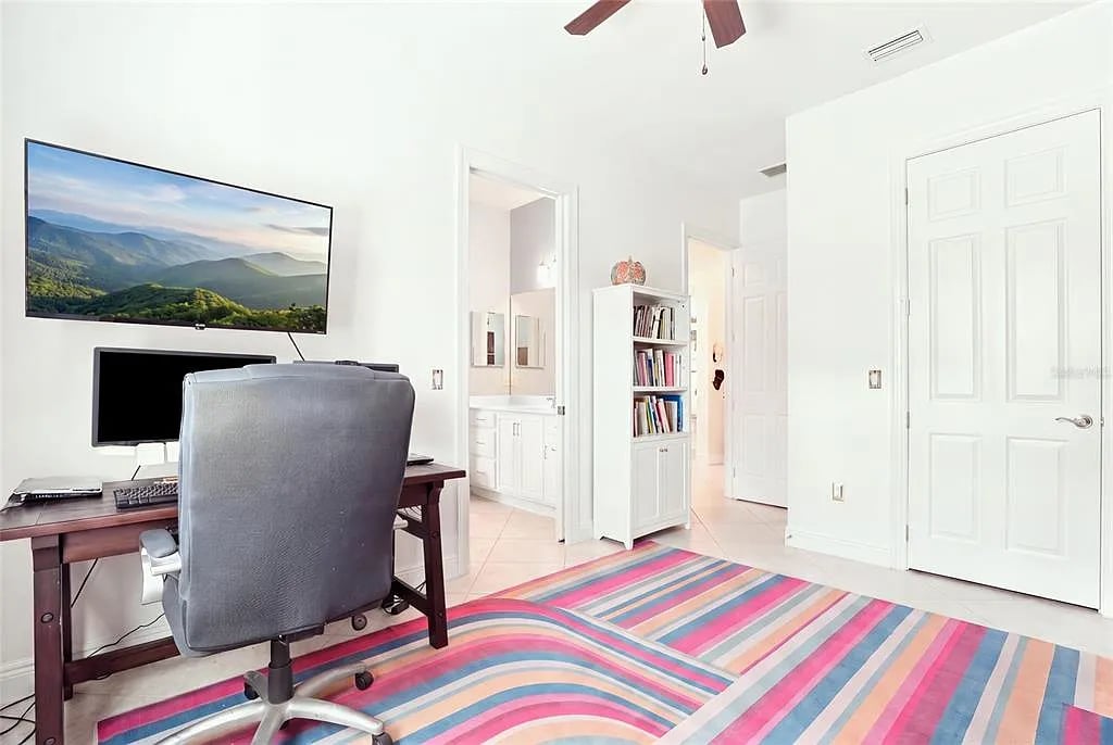 Bright home office with a dark desk, wall-mounted television, white bookcase, and colorful striped rug.