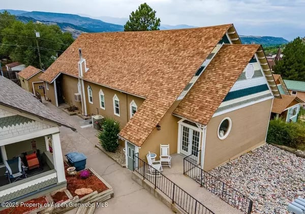 Tan residential building with steep shingle roofs and a paved walkway, with mountains in the background.