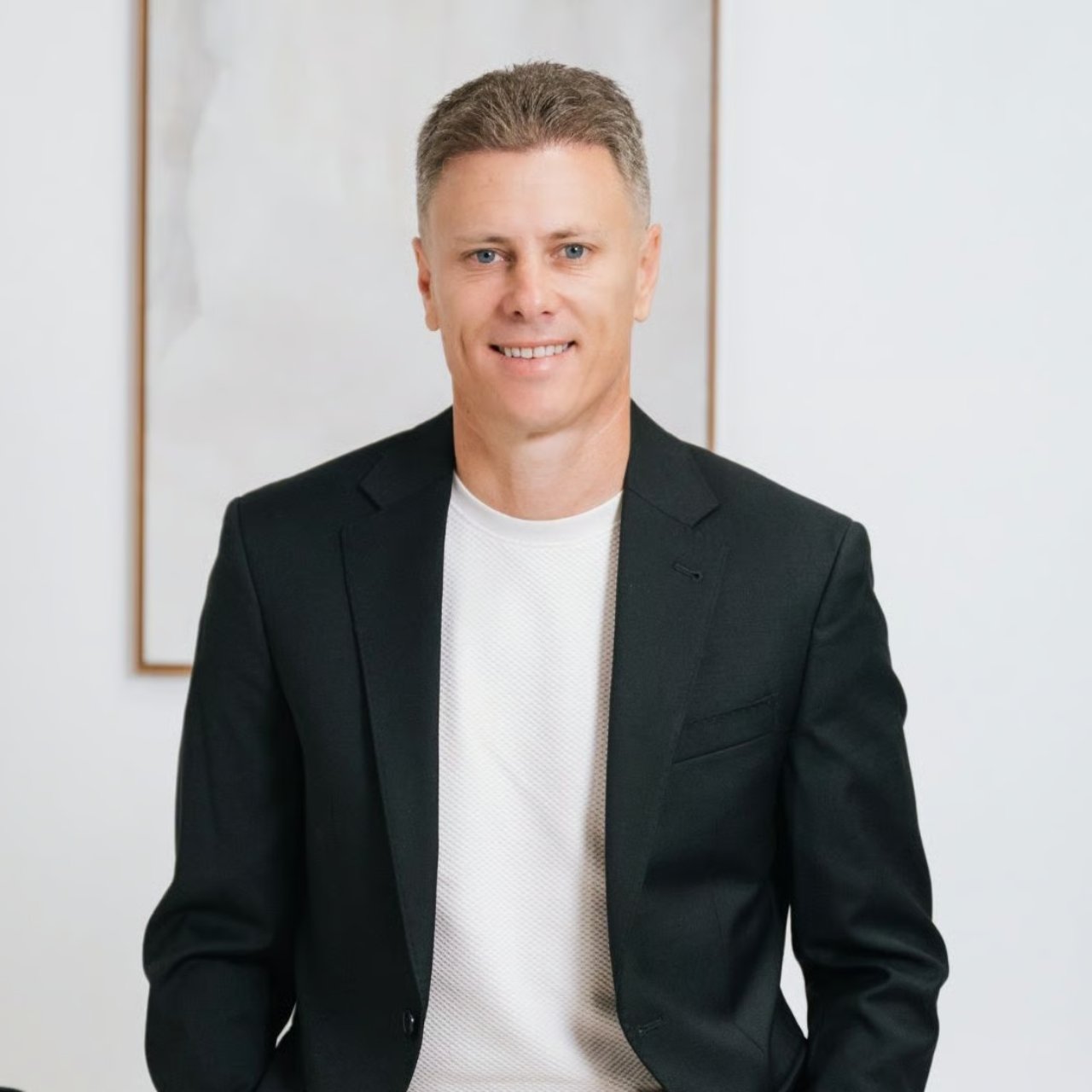 Smiling man in a black blazer and white shirt against a bright indoor backdrop.