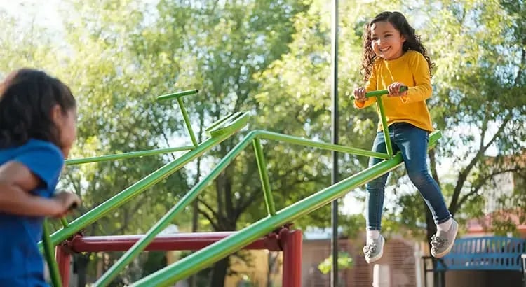 Two children are seen playing on a green playground structure. The scene captures the joy and energy of outdoor playtime.