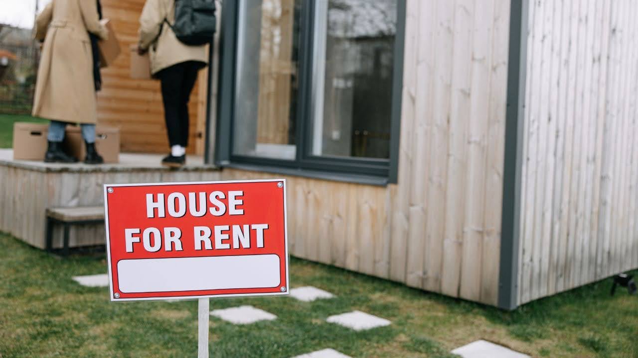 a-red-house-for-rent-sign-stands-on-a-lawn-in-front-of-a-modern-wooden-house-in-the-background-two-people-enter-the-house-carrying-boxes