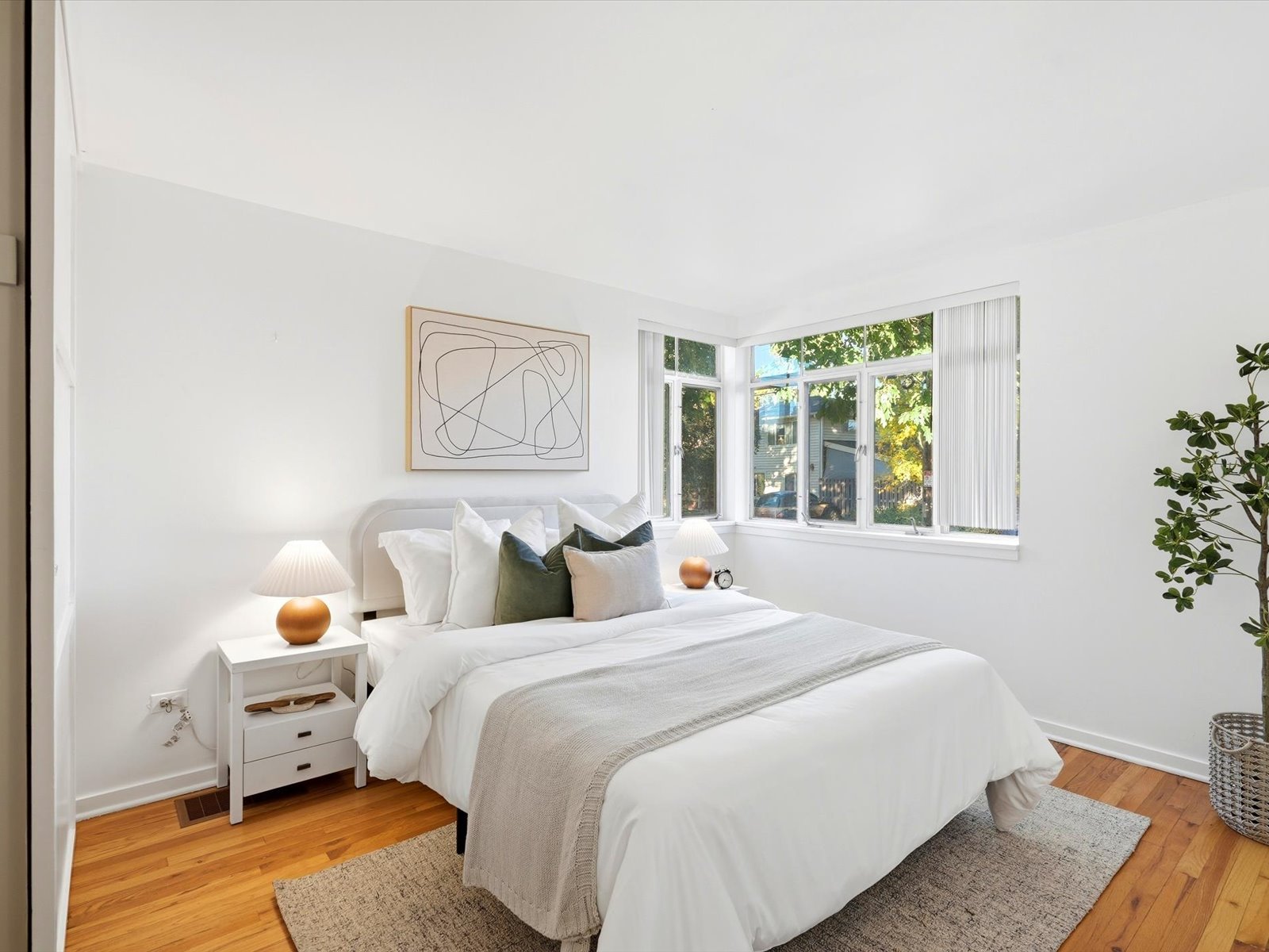 Staged bedroom with a white bed, large corner windows, and warm hardwood floors.