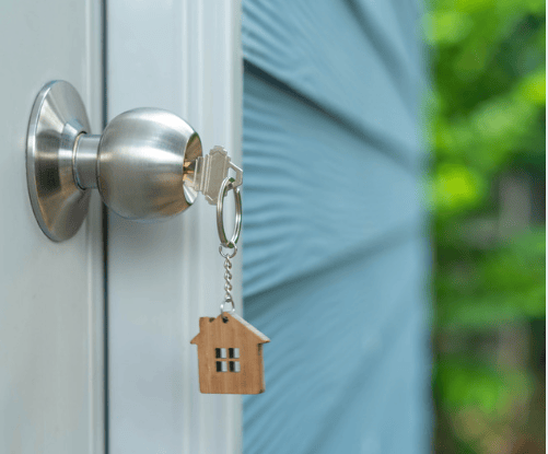 Keys with a wooden house-shaped keychain hang from a silver doorknob beside blue siding.