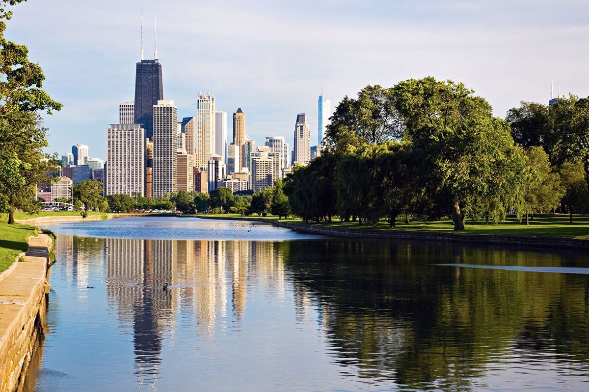 Chicago skyline reflected in a park lagoon, with Willis Tower rising above the trees.