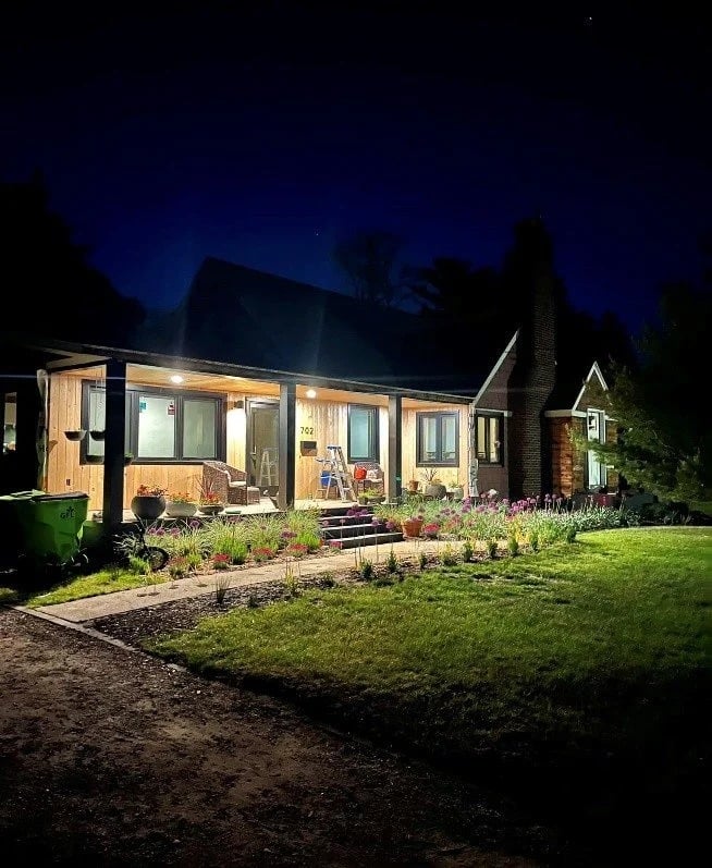 Renovated house exterior at night, with ladders on the porch and flower beds along the walkway.