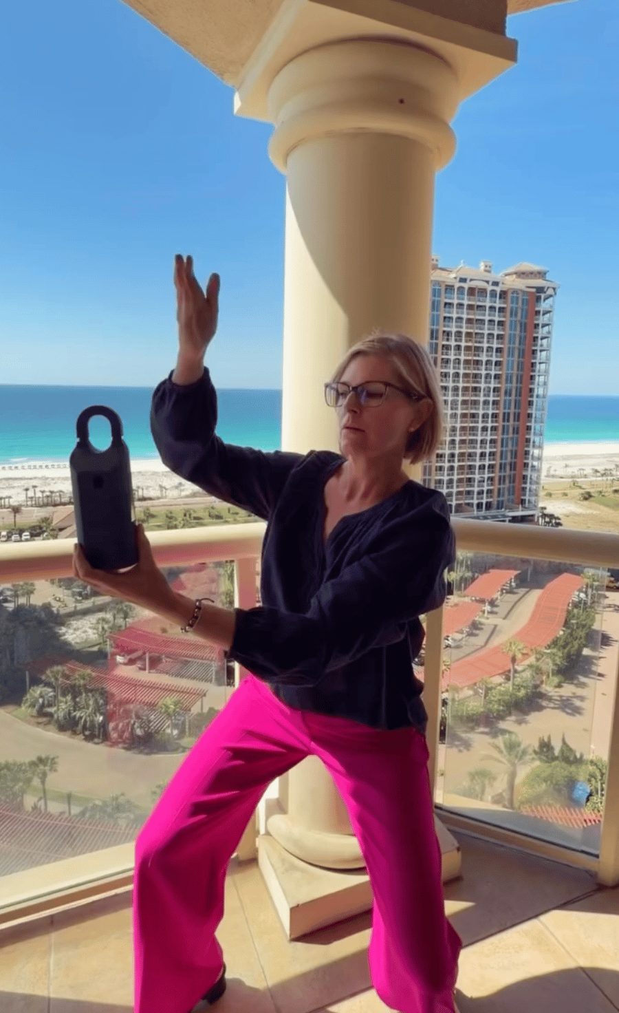 Woman posing with a portable speaker on a balcony overlooking the beach and ocean.