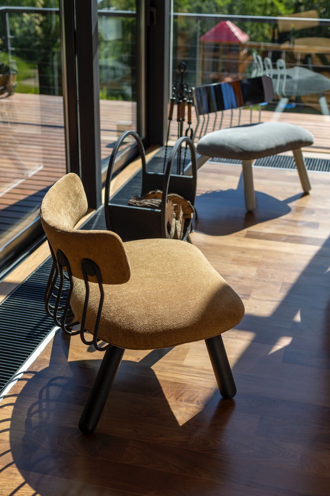 Ochre upholstered chair beside a firewood holder and glass doors opening onto a timber deck.