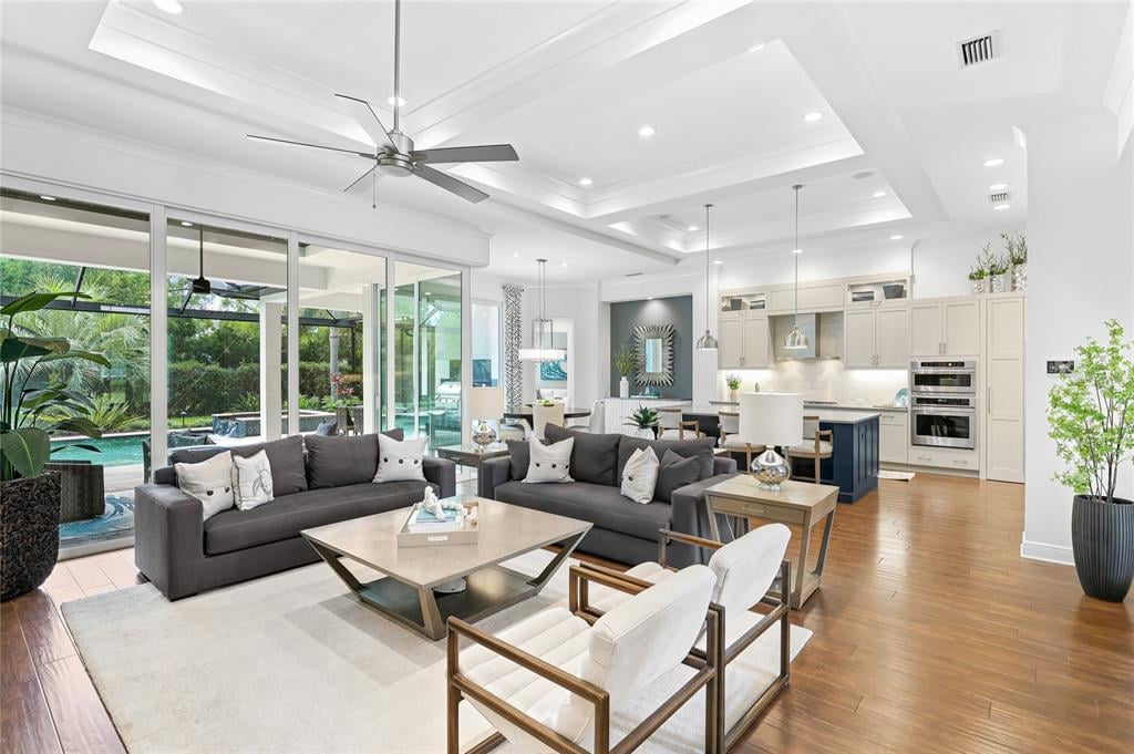 Open-plan living room and kitchen with gray sofas and sliding doors overlooking a pool and tropical landscaping.