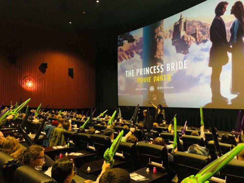 Movie Magic: At an Alamo Drafthouse Movie Party, fans of The Princess Bride fill the theater, waving green props in excitement as the screen lights up with the film’s iconic romance, creating a lively, interactive vibe.