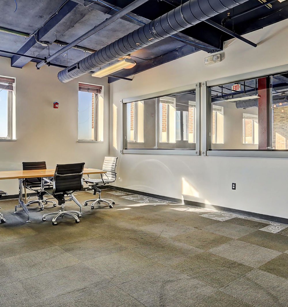 Office meeting room with a conference table, rolling chairs, and exposed ceiling ductwork.