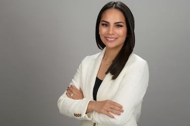 Woman in a white blazer with straight dark hair, smiling with her arms crossed.