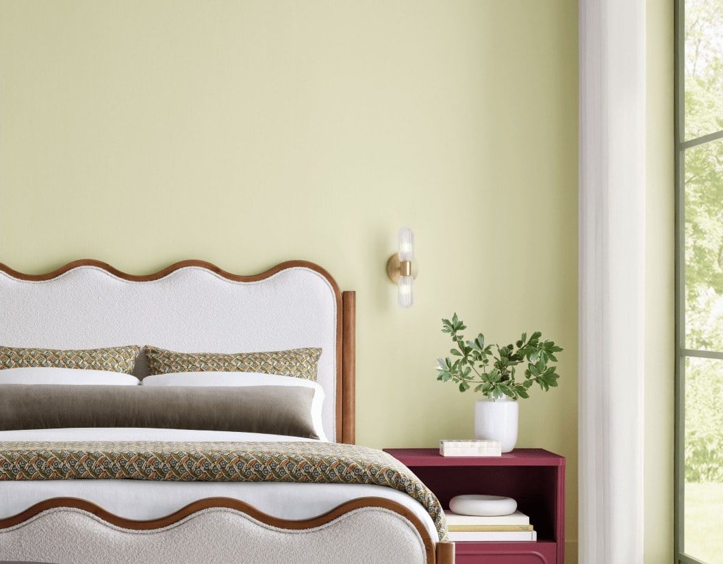 A bedroom with pale yellow-green walls painted Celery SW 6421, a white upholstered bed with a scalloped wood frame, and a deep red nightstand.