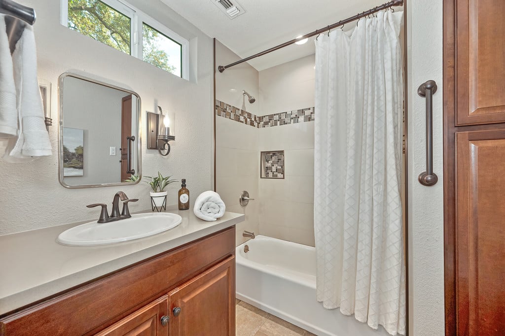 Bathroom with a wood vanity, oval sink, and curtained bathtub beneath high windows.