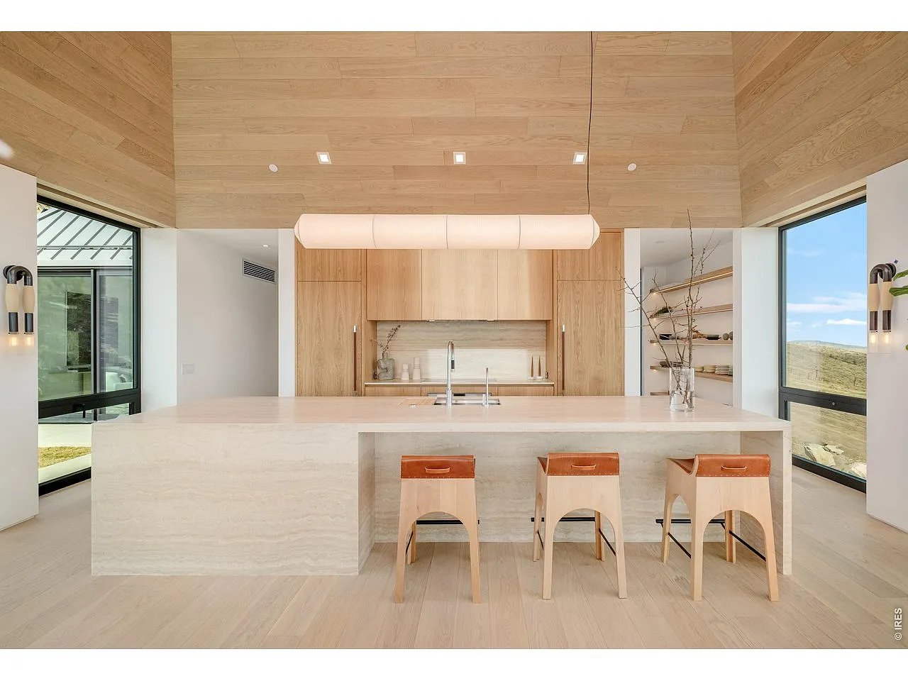 Modern kitchen with a pale stone island, three stools, oak cabinetry, and views of rolling hills.